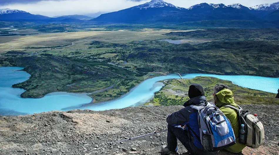 Two hikers with backpacks overlooking a sinuous blue river down in a valley with mountains in background